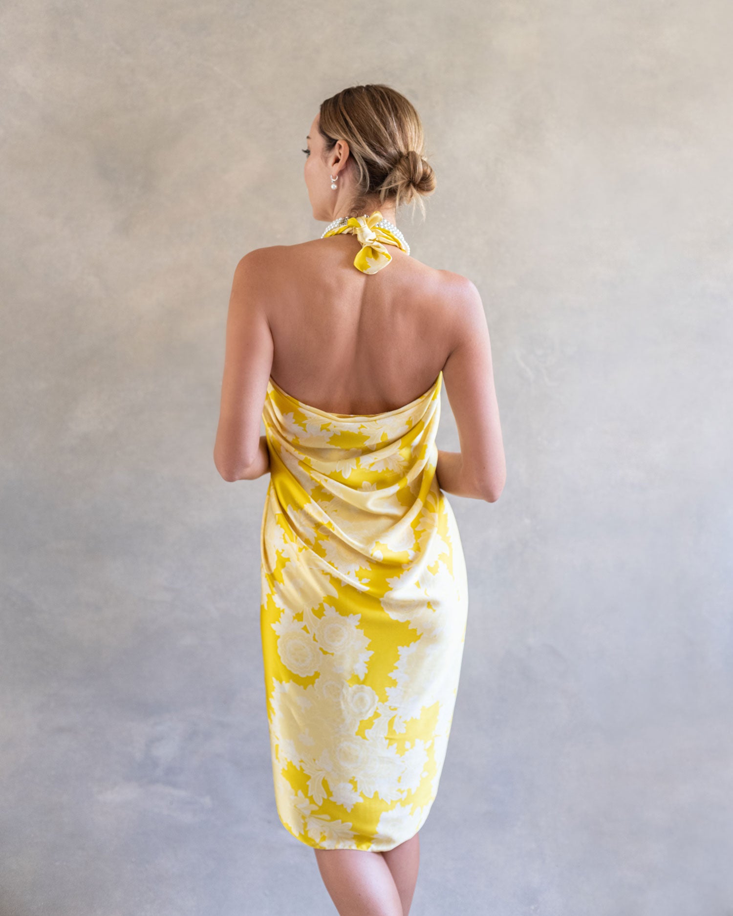 Elegant back view of a woman wearing a yellow sarong pareo styled as a dress, the colorful peacock and flower design vivid against a neutral studio backdrop