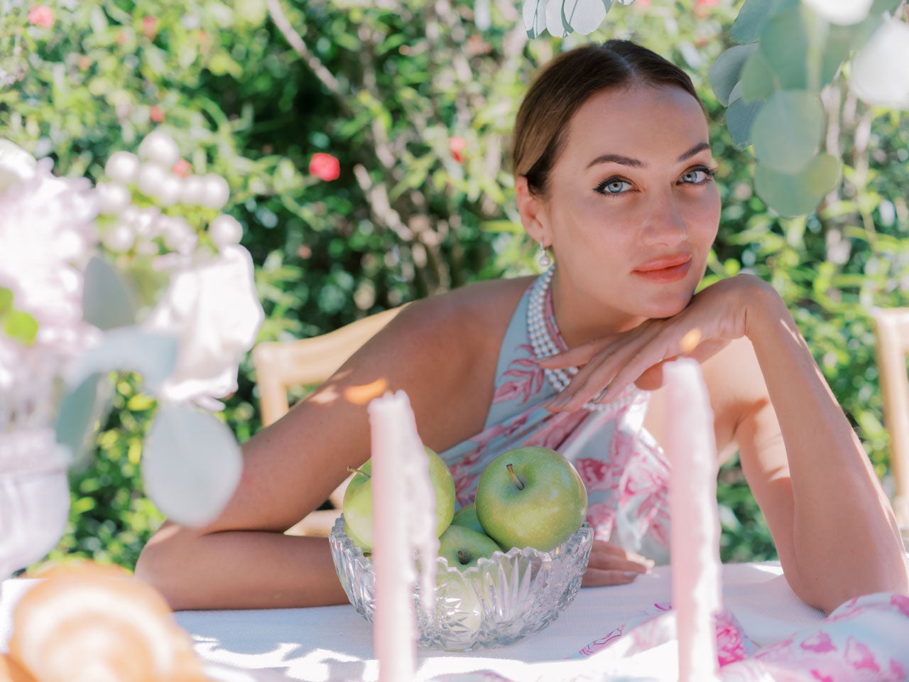 Serene image of woman sitting at table with candles, garden in background, wearing pink and blue pareo dress and pearls