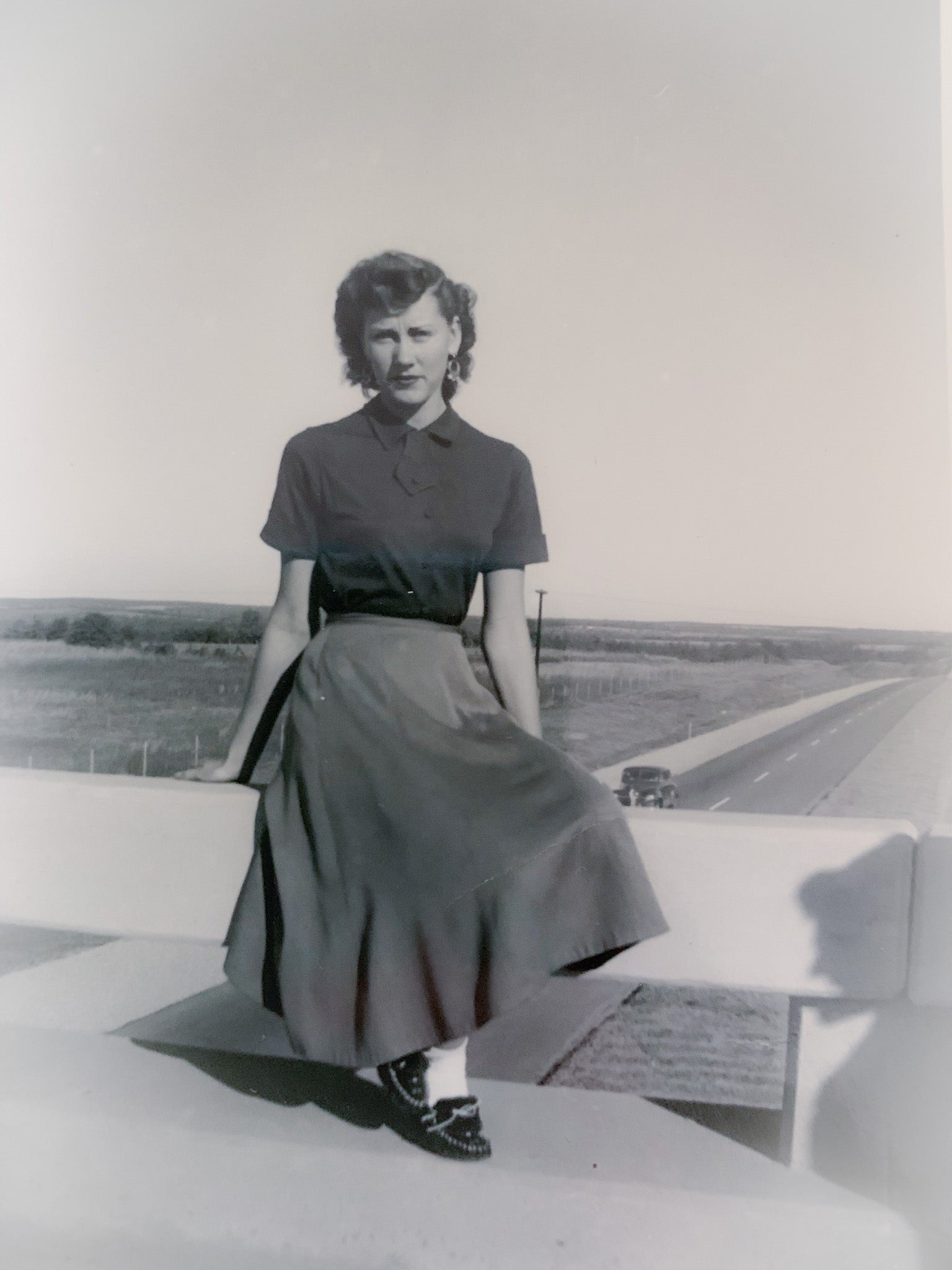 Black and white historical photo of Virgie Wayne from the 1950s, depicting her seated gracefully on a highway overpass in Texas, dressed in a classic 1950s skirt and shirt, embodying the era's style