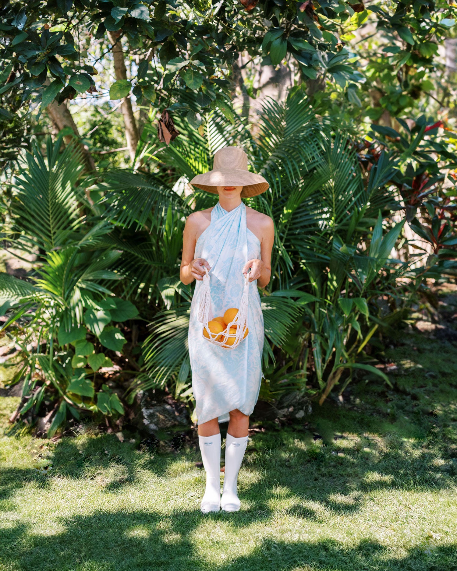 Playful and artistic shot of woman in a light blue sarong dress and retro hat, creating a sophisticated and timeless garden look as she holds fresh oranges against a lush palm background, evoking a sense of summer abundance