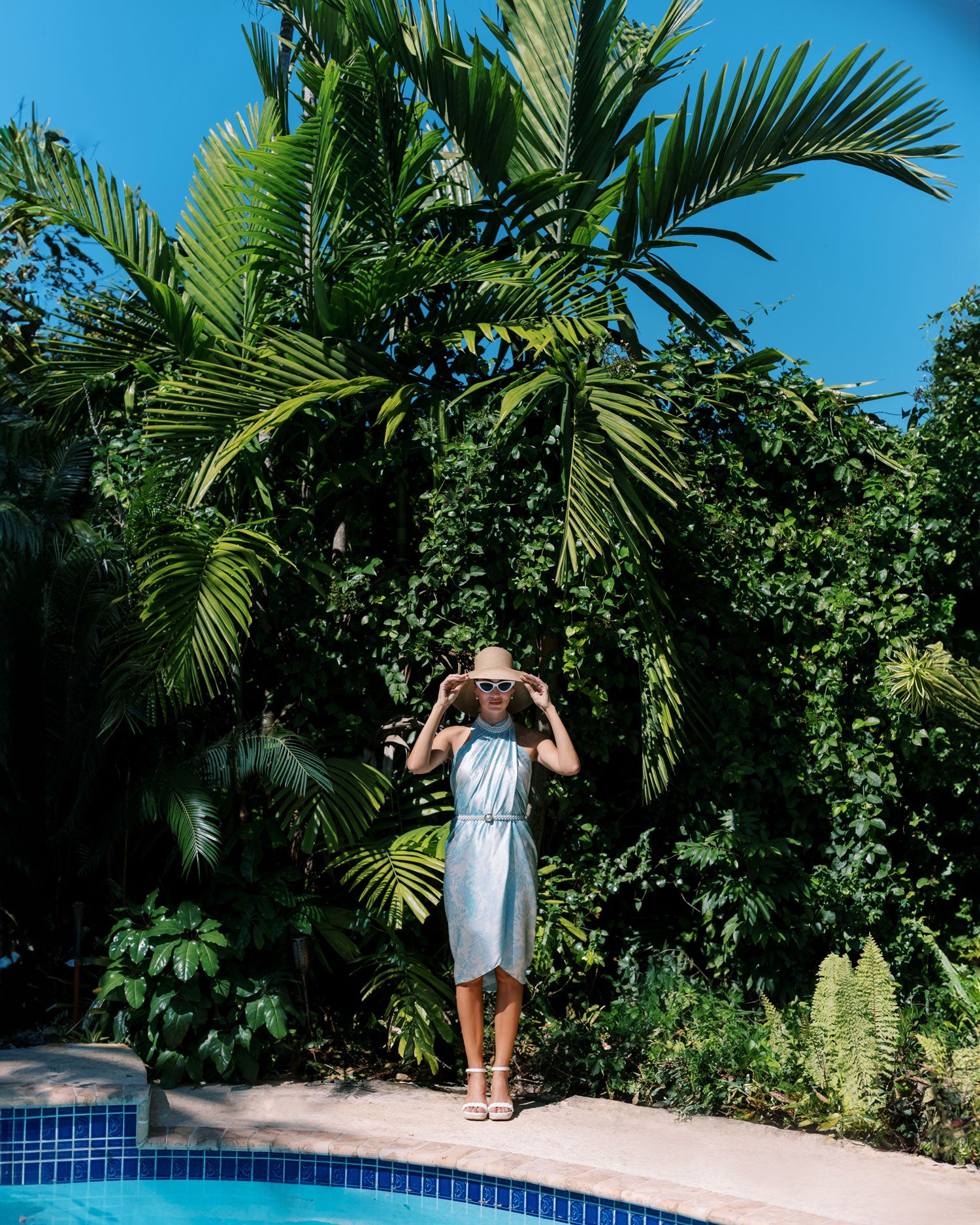 Playful and stylish image of a woman in a light blue pareo dress in retro sunglasses and hat against and lush tropical background and next to a bright blue pool