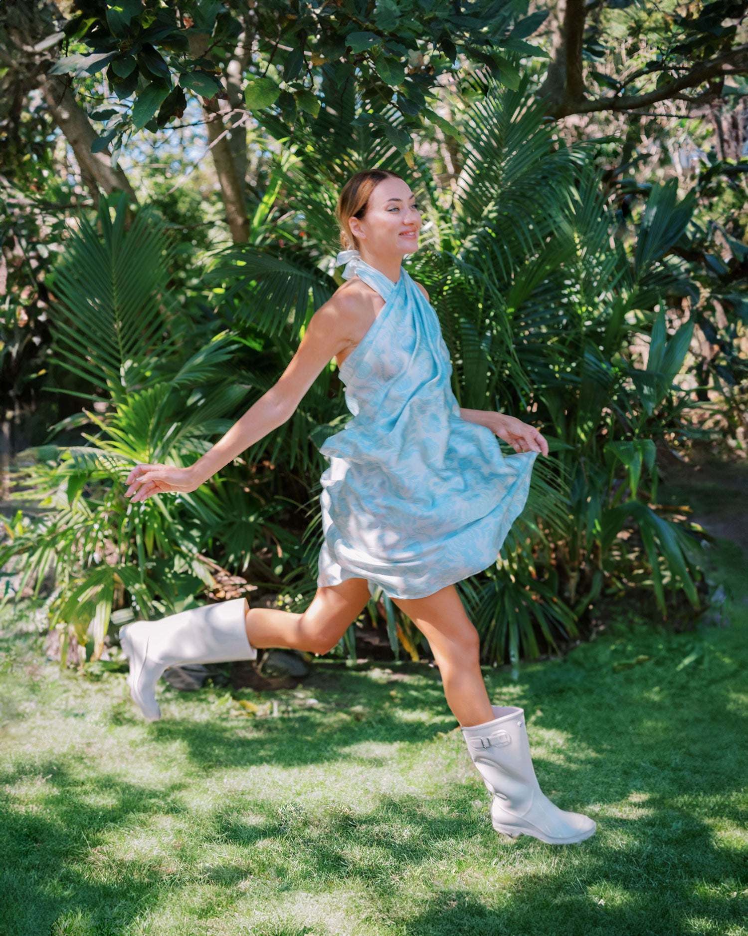 Joyful woman in a garden, her sky blue sarong pareo dress capturing the essence of playful elegance as she jumps, the fabric billowing around her