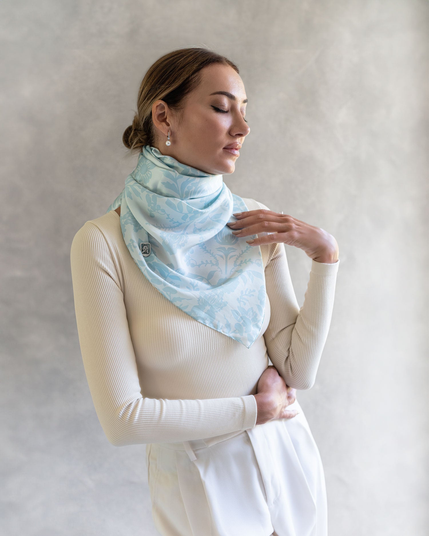 Woman wearing a sky blue pareo as a sophisticated scarf, the fabric softly enveloping her eyes are closed in a moment of reflection, soft studio background