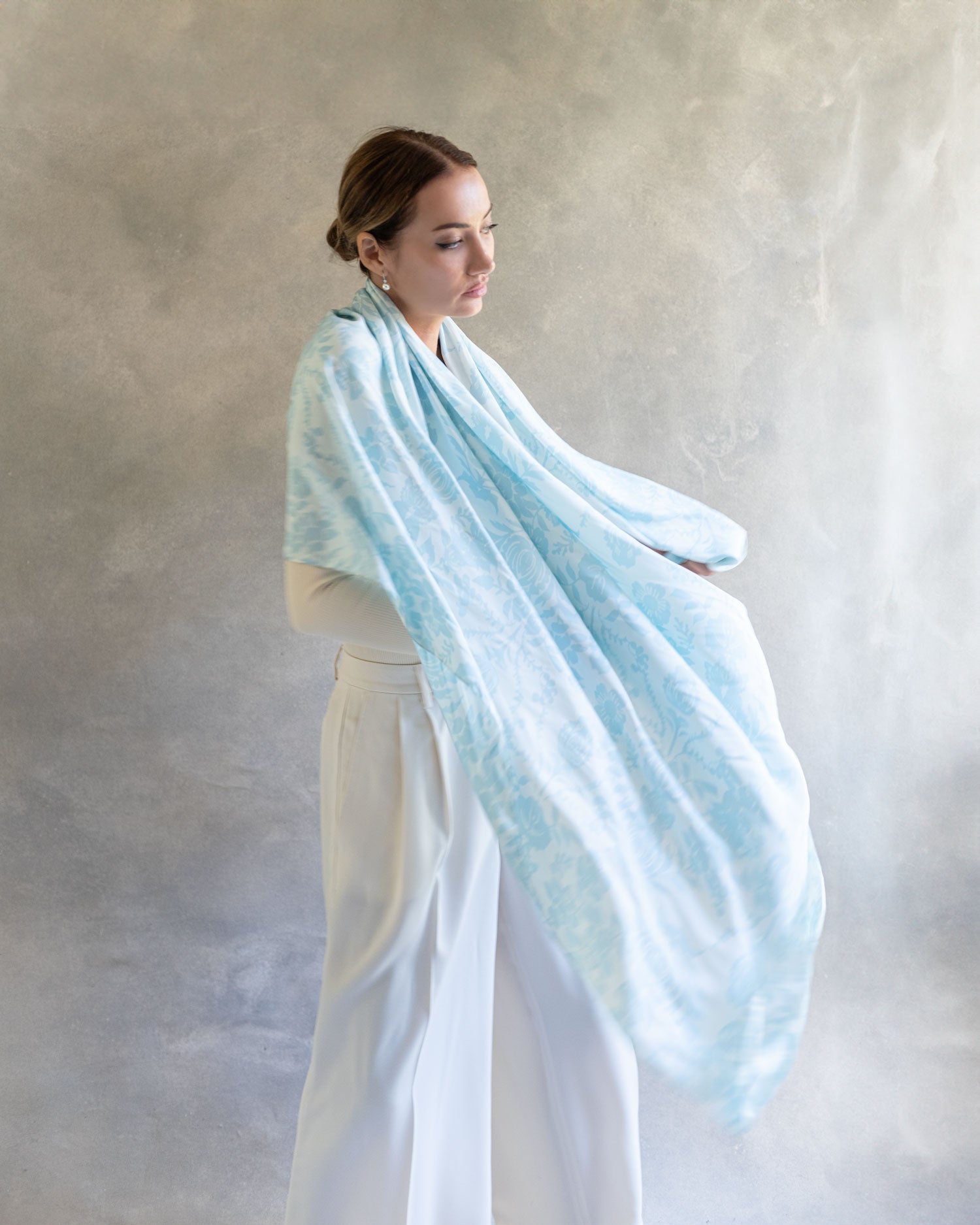 Woman draped in a light sky blue pareo used as a shawl, exuding elegance and versatility in a tranquil indoor studio setting