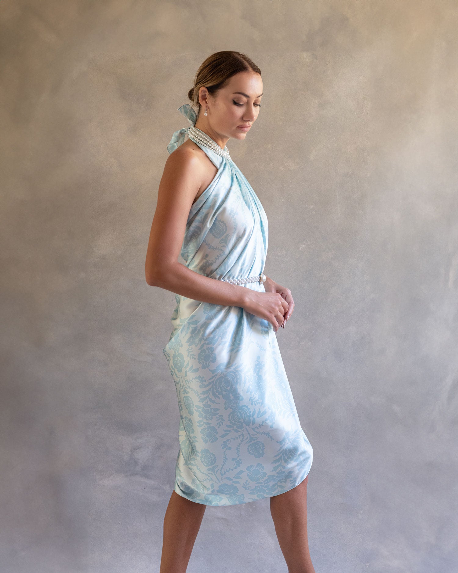 Side profile of a woman in a sky blue pareo dress, highlighting its delicate drape and ethereal quality against a soft background