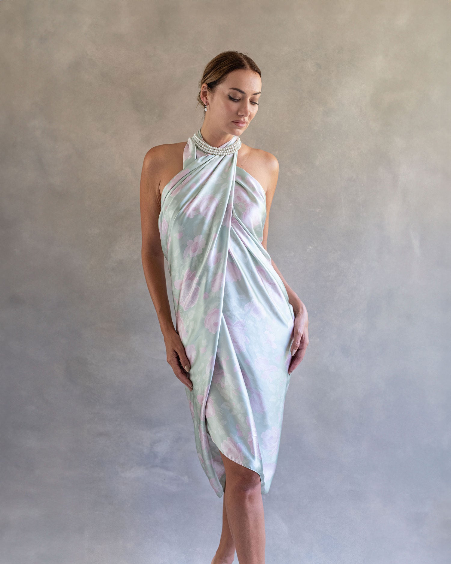 Front view of a woman in a light jade green pareo wrap dress, adorned with a delicate pastel pink rose pattern, standing against a serene grey studio backdrop.