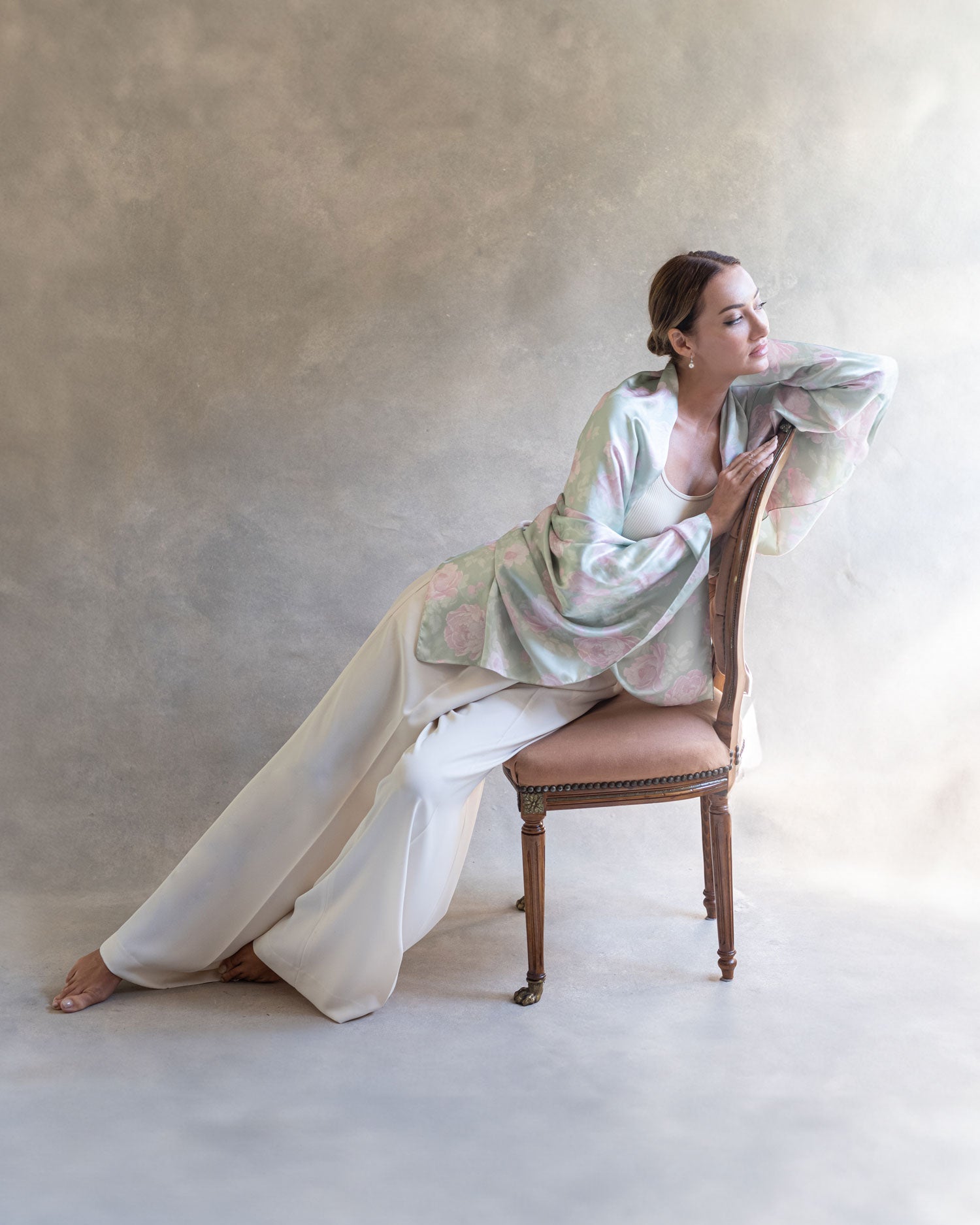 Full body shot of a woman seated in an elegant antique chair, wrapped in a jade green pareo dress, looking out a window in a thoughtful pose