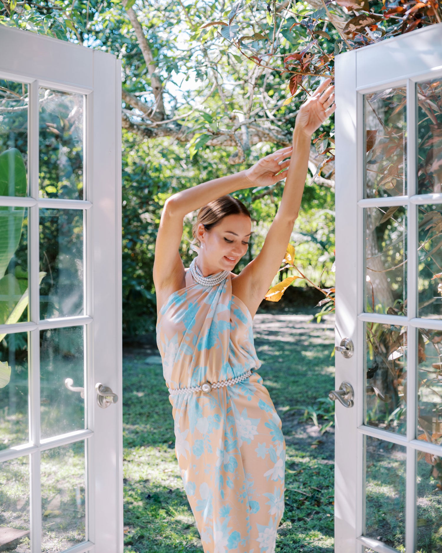 Playful front view of a woman posing in an orange and blue sarong pareo, styled as a dress, between two open french doors with tropical setting in background