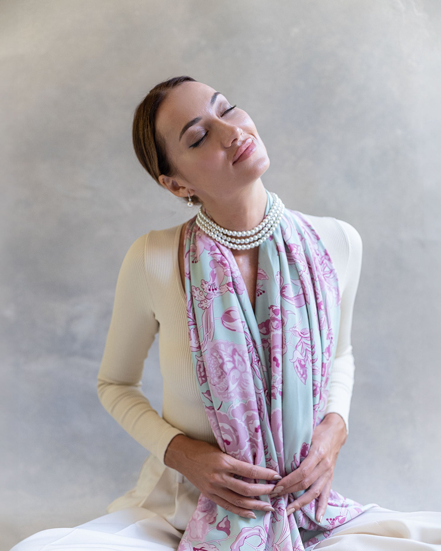 Fashion model seated in a light pink and blue silk pattern scarf, front view, serene and happy with eyes closed against soft grey background