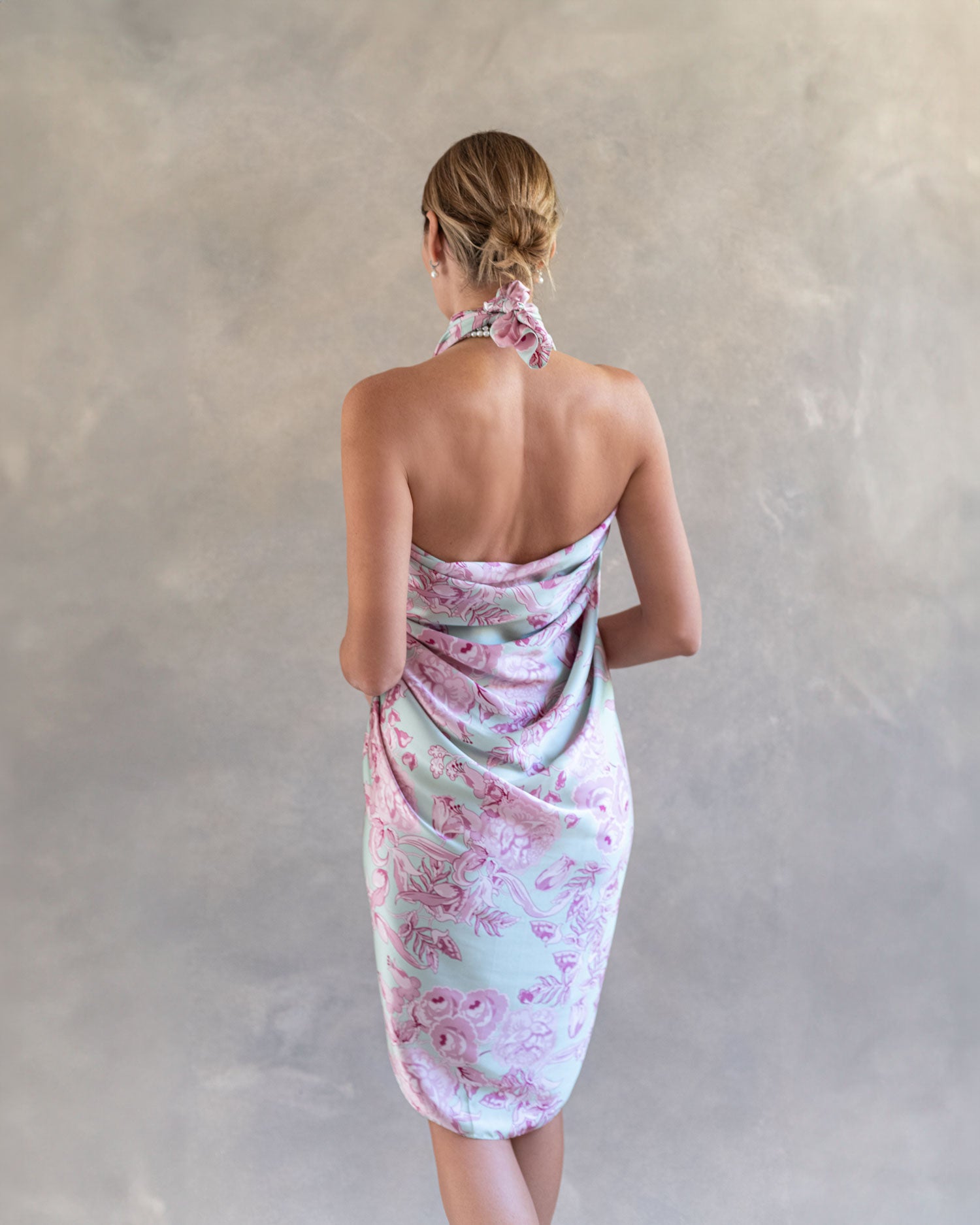 Fashion model posing in a light pink and blue silk pareo, back view, styled as a sophisticated dress against soft grey background