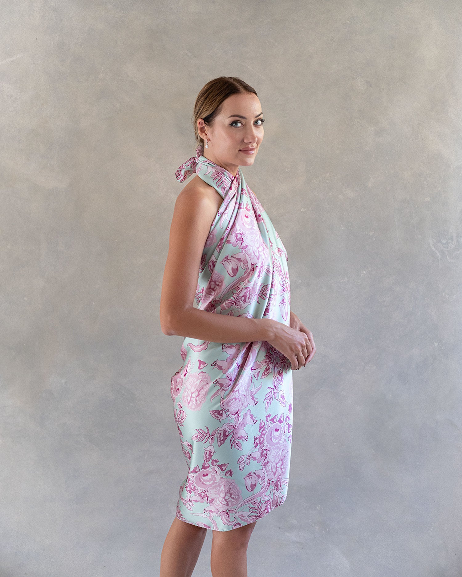 Fashion model posing in a light pink and blue silk pareo, side view, styled as a sophisticated dress against soft grey background