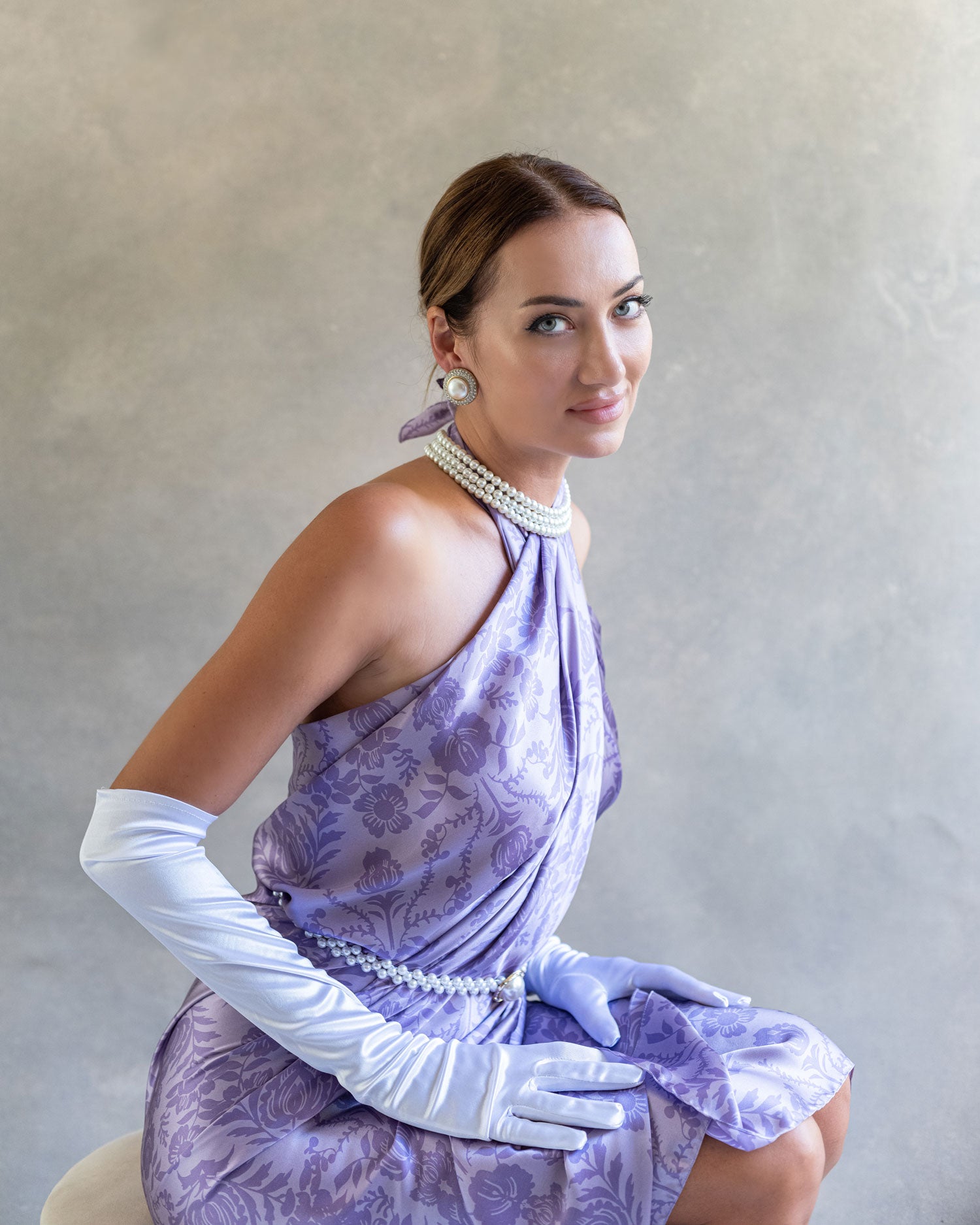 Elegant side view of seated woman showcasing a purple lavender sarong pareo dress, highlighting its elegance, wearing pearls and long white gloves