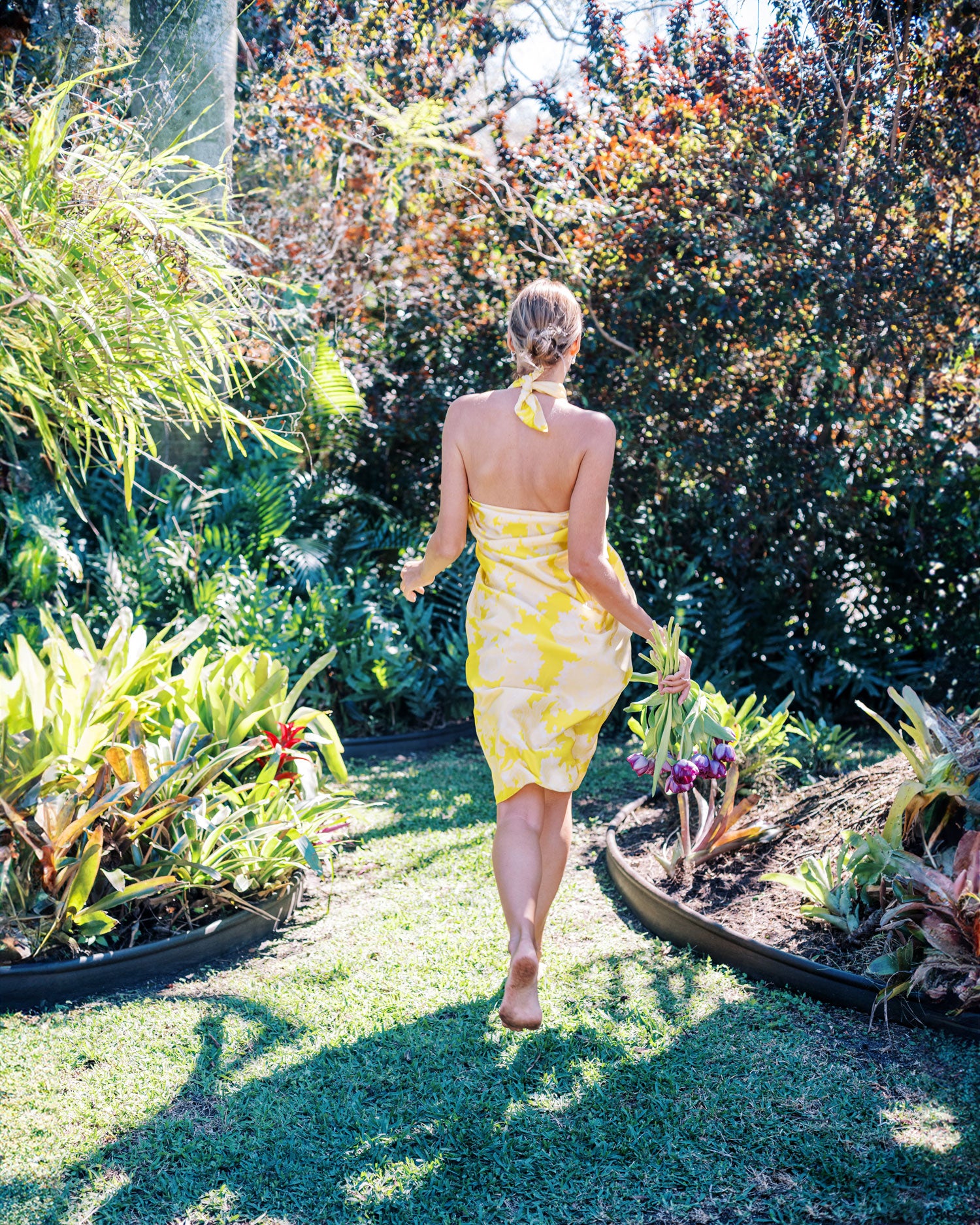 Back view of a woman running through tropical garden holding purple flowers, her bright yellow pareo dress with peacock pattern fluttering in the breeze