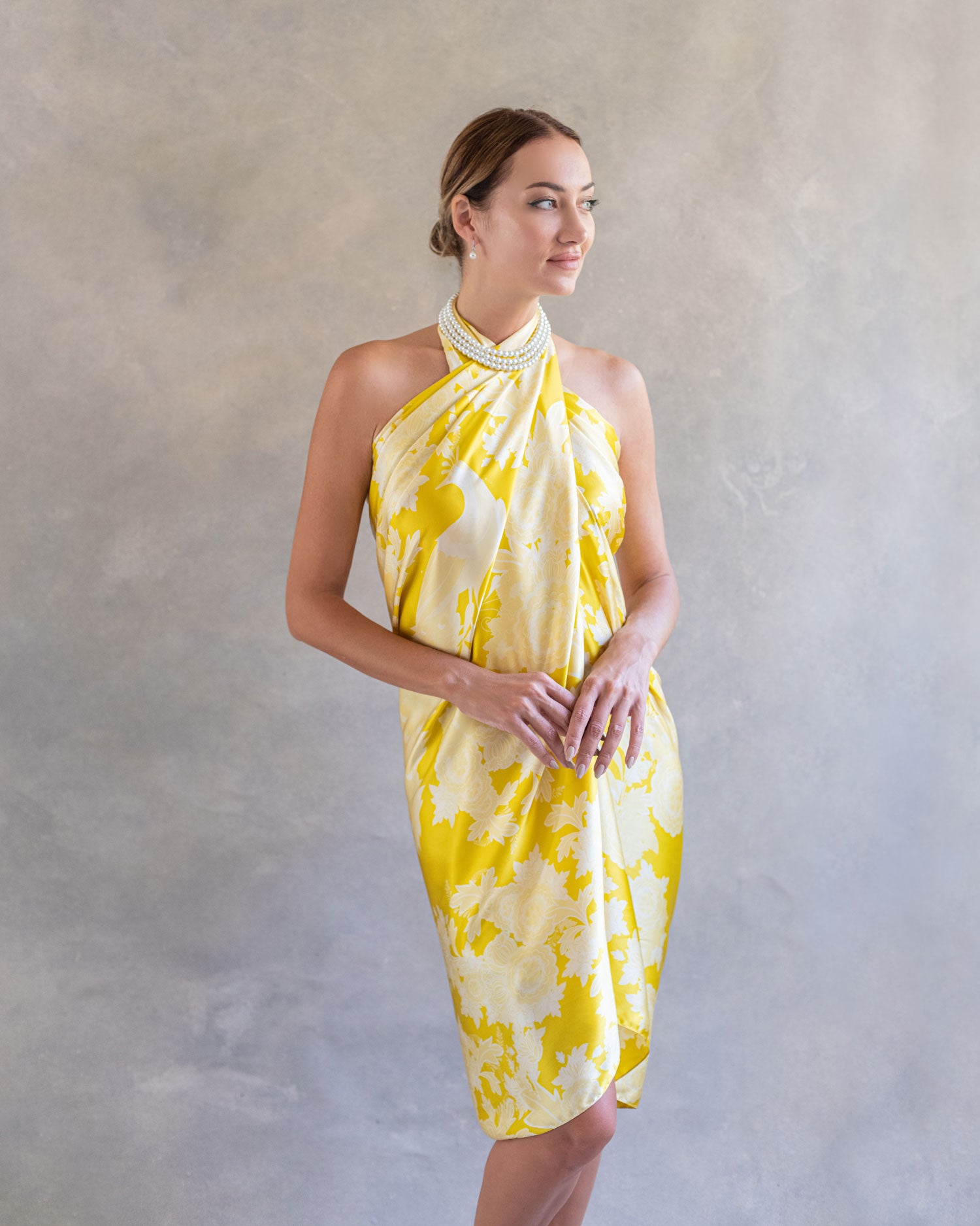 Front view of a woman in a bright yellow sarong pareo wrap dress adorned with a peacock and flower pattern, standing elegantly in a serene studio setting