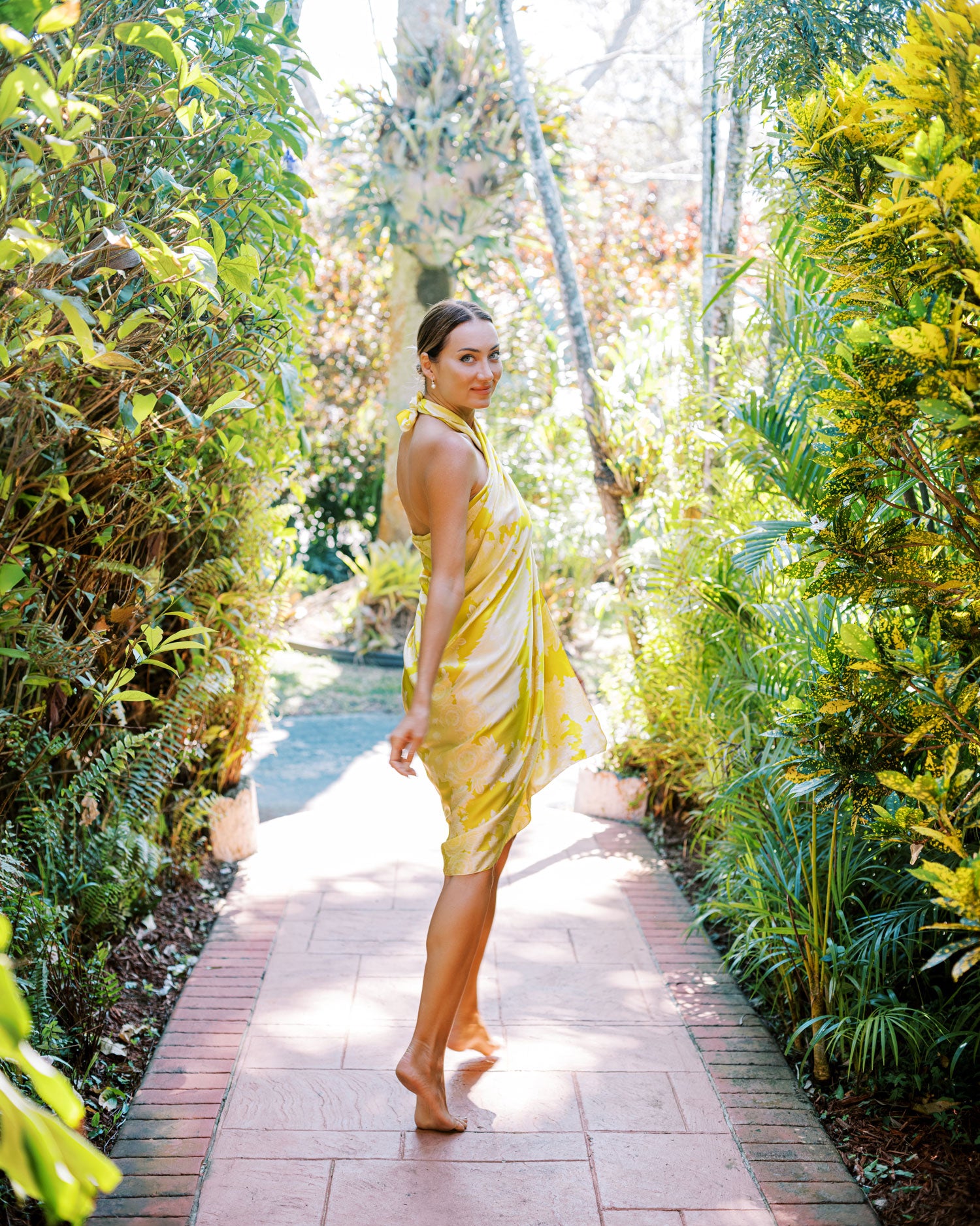 Woman walking between hedges and palm trees, her bright yellow pareo sarong dress flowing gracefully, mirroring the lush greenery around her