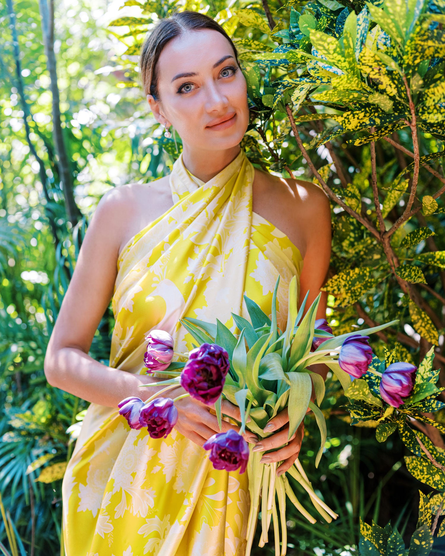 Close-up of a woman holding purple flowers, making eye contact with the camera, her bright yellow pareo adorned with peacock and flower motifs framing her face