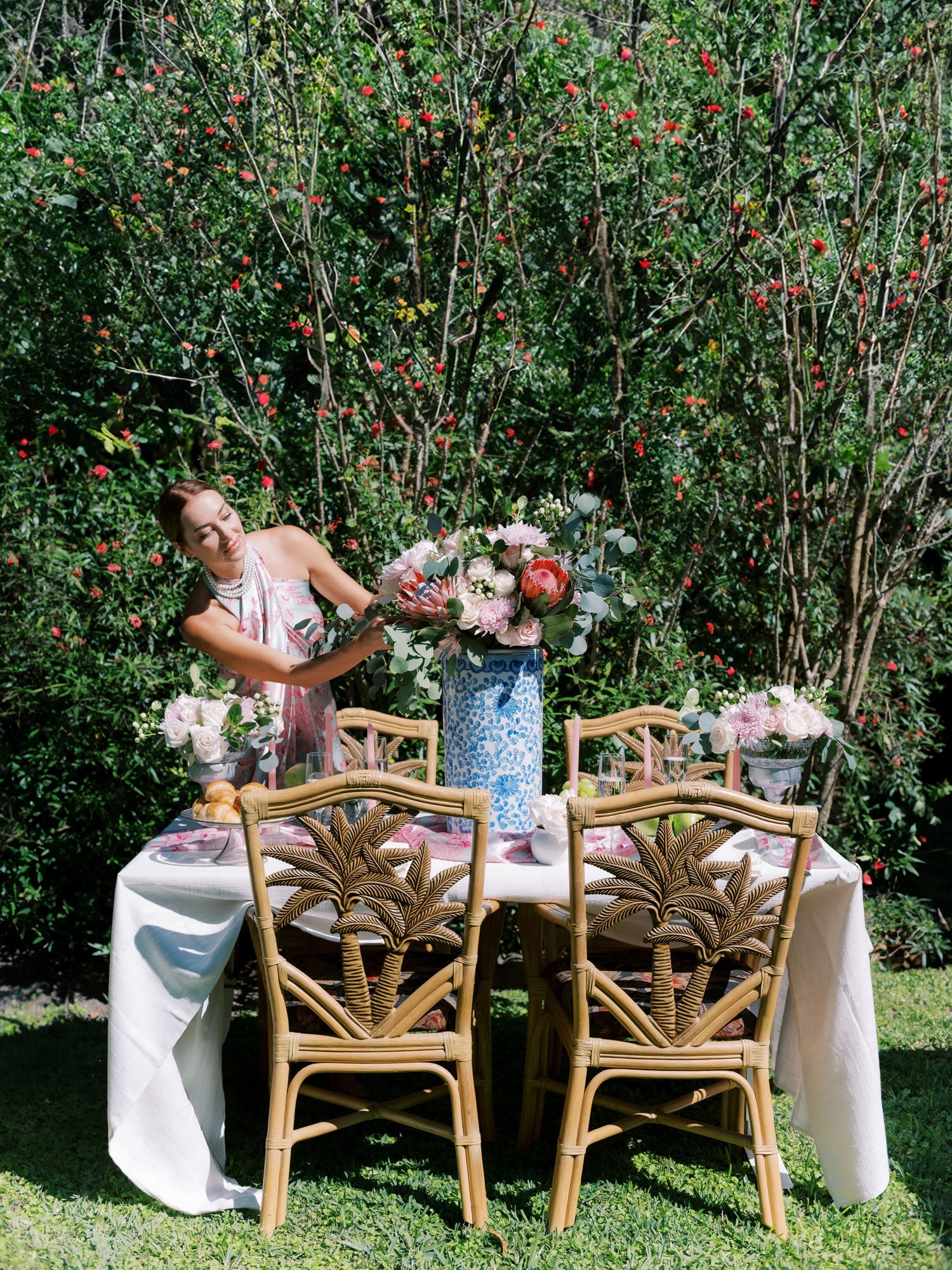 Woman in a pink and blue pareo dress arranging a bouquet at an outdoor table, with lush pink flowers blooming in the background, enhancing the vibrant, floral setting