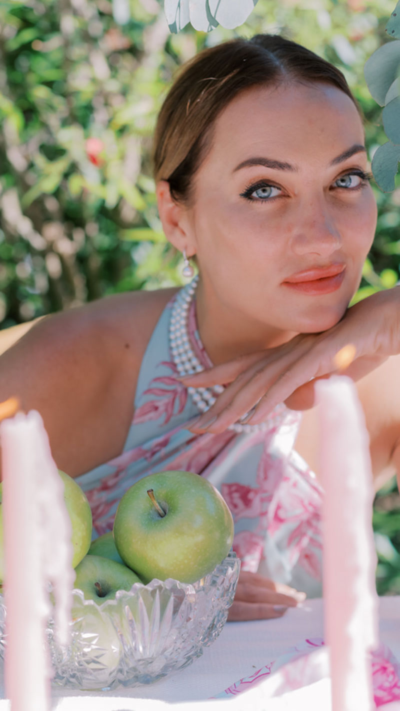 Close up of woman sitting at table with candles and green apples, garden in background, wearing pink and blue pareo dress and pearls