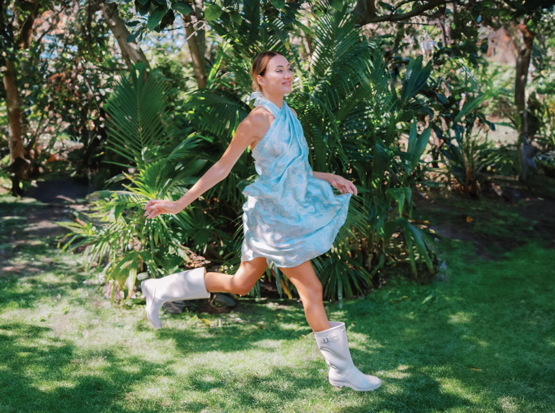 Joyful scene of a woman in a garden, her sky blue pareo dress capturing the essence of playful elegance as she jumps, the fabric billowing around her